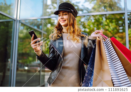 Smiling woman with shopping bags using her phone. Young woman after shopping on the city street. Smiling woman with shopping bags using her phone. Young woman after shopping on the city street. 111891625