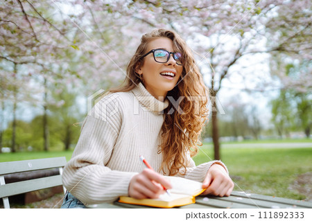 Young woman sitting at a table in blooming spring park with a notepad and laptop. Lifestyle concept. 111892333