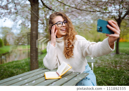Selfie time. Young woman takes selfie photo in blooming spring park. Concept of relaxation, walking. Selfie time. Young woman takes selfie photo in blooming spring park. Concept of relaxation, walking. 111892335