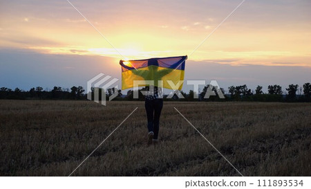 Happy lady walking on barley meadow and turning around with a raised over head flag of Ukraine. Ukrainian woman with a lifted blue-yellow banner on a beautiful sunset at background. End of war. 111893534