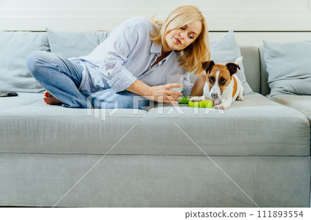 Lovely charming mature mid adult blond female play, caressing and pet dog while sitting at cozy living rom at home interior. Lovely charming mature mid adult blond female play, caressing and pet dog while sitting at cozy living rom at home interior. 111893554