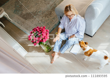 A bouquet of roses in a glass vase nearby adult beautiful female people - enjoying quiet lifestyle indoor caressing playing with her adorable jack Russell terrier. 111893595