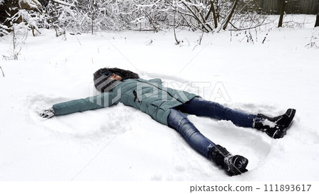 Happy woman lying on snowy lawn at forest and making angel. Young girl playing in snow and enjoying wintertime. Cheerful lady having fun outdoor. Concept of a winter vacation. 111893617