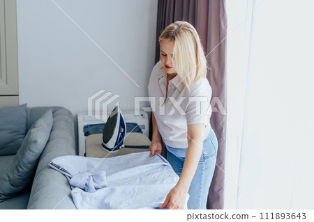 Mature blond woman ironing her blouse before going to walk at cozy home interior. Modern female ironing clothes after doing laundry, using heat and steam to iron clothing on board. 111893643