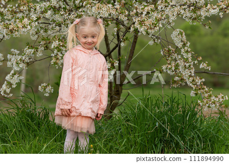 Surprised fair-haired girl with two ponytails in light pink dress stands near flowering tree. Surprised look with raised eyebrows Surprised fair-haired girl with two ponytails in light pink dress stands near flowering tree. Surprised look with raised eyebrows 111894990