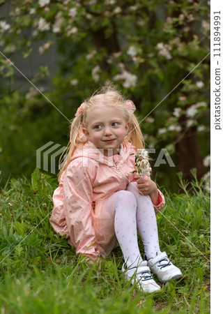 Smiling girl with two blond hair in pink jacket with flowering branch in hand sits on grass. Blossom tree background. Springtime 111894991
