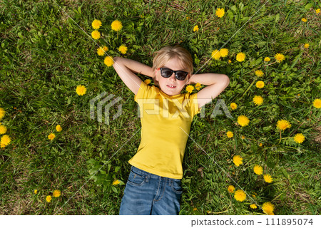 Happy boy puts his hands behind his head. Blond child lies on grass in dandelions field and dreaming. Top view 111895074