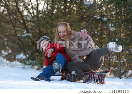 Young mother and teenage son fool around in woods in winter. Woman and boy have picnic in snowy forest. Young mother and teenage son fool around in woods in winter. Woman and boy have picnic in snowy forest. 111895113