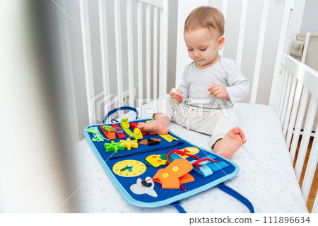 Baby playing with montessori busy book sitting in crib. Educational books and quiet books concept. Montessori busy board Baby playing with montessori busy book sitting in crib. Educational books and quiet books concept. Montessori busy board 111896634