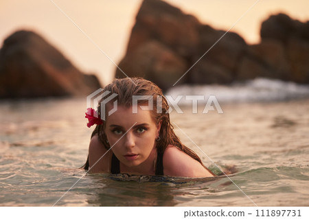 Woman with red flower in hair swims in serene rocky lagoon. Female enjoys tranquil water in picturesque nature spot. Travel leisure, exotic destination, adventure seeker in natural setting. 111897731