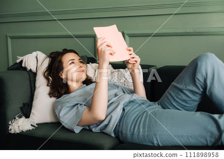 Contented Woman Relaxing on Sofa with Book, Enjoying Leisure Time. Smiling Young Lady Engaged in 111898958