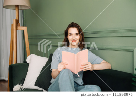 Happy young woman reading a book sitting comfortably on the couch enjoying a relaxing evening 111898975