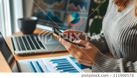 Young business woman sitting in office at table and using smartphone. On desk is laptop and tablet computer, on screen charts and graphs. Woman analyzing data. Student learning online. Young business woman sitting in office at table and using smartphone. On desk is laptop and tablet computer, on screen charts and graphs. Woman analyzing data. Student learning online. 111900578
