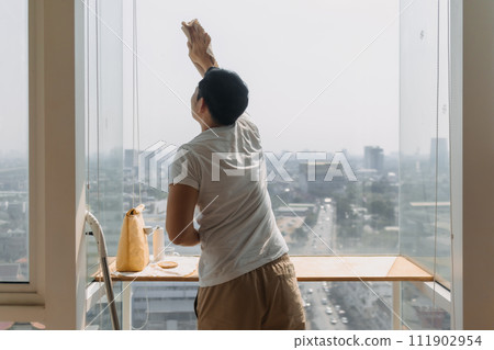 Backside view of asian Thai man wiping window glass in room apartment with city view, keep glass sparking clean, house husband and housework concept. 111902954