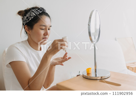 Asian Thai woman using and applying sun cream moisturizer in front of the mirror while sitting on white couch in room apartment, beauty concept. 111902956