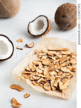 Dried organic coconut slices chips on baking paper with whole ripe coconut on light background. Dried organic coconut slices chips on baking paper with whole ripe coconut on light background. 111907361