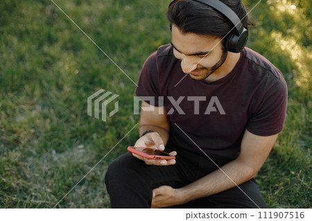 Young boy listening to music while sitting on a grass. Man using a smartphone. Man wearing a bordo shirt and jeans. Young boy listening to music while sitting on a grass. Man using a smartphone. Man wearing a bordo shirt and jeans. 111907516