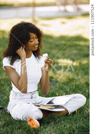 African girl holding a earpods while sitting on a grass. Girl is using a white tablet. Girl wearing a white clothes. African girl holding a earpods while sitting on a grass. Girl is using a white tablet. Girl wearing a white clothes. 111907524