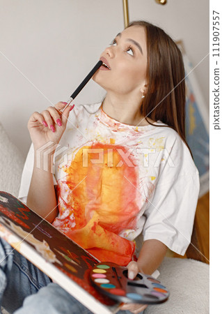 Young female artist posing holding brushes and painting in hands. Woman wearing white t-shirt and blue jeans. Brunette girl sitting in a chair in room with white wall. 111907557