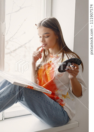 Young female artist posing holding brushes and painting in hands. Woman wearing white t-shirt and blue jeans. Brunette girl sitting on a windowsill in room with white wall. Young female artist posing holding brushes and painting in hands. Woman wearing white t-shirt and blue jeans. Brunette girl sitting on a windowsill in room with white wall. 111907574
