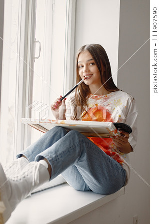 Young female artist posing holding brushes and painting in hands. Woman wearing white t-shirt and blue jeans. Brunette girl sitting on a windowsill in room with white wall. 111907590