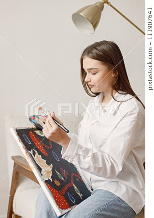 Satisfied female artist posing holding new picture and paint palette in hands. Woman wearing jeans and white shirt. Brunette girl sitting on a chair in room with white wall. Satisfied female artist posing holding new picture and paint palette in hands. Woman wearing jeans and white shirt. Brunette girl sitting on a chair in room with white wall. 111907641