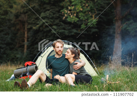 Man and woman with backpacks sitting on a grass in forest near fire. Couple lighting a fire near tent in forest at summer during their trip. Man and woman wearing green t-shirts. Man and woman with backpacks sitting on a grass in forest near fire. Couple lighting a fire near tent in forest at summer during their trip. Man and woman wearing green t-shirts. 111907702