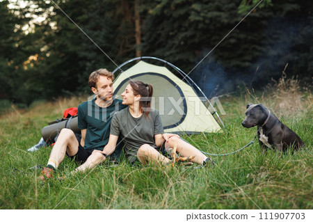 Man and woman with backpacks sitting on a grass in forest near fire. Couple lighting a fire near tent in forest at summer during their trip. Man and woman wearing green t-shirts. Man and woman with backpacks sitting on a grass in forest near fire. Couple lighting a fire near tent in forest at summer during their trip. Man and woman wearing green t-shirts. 111907703