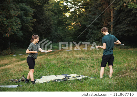 Man and woman with backpacks on a grass in forest and setting up a tent. Couple with a tent in forest at summer during their trip. Man and woman wearing green t-shirts. 111907710