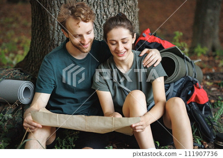 Couple with backpacks sitting on a grass in forest and looking in a map. Man and woman resting in forest at summer during their trip. Man and woman wearing green t-shirts. 111907736