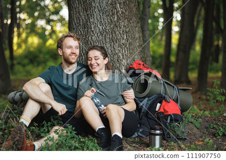 Couple with backpacks sitting on a grass in forest. Man and woman resting in forest at summer during their trip. Man and woman wearing green t-shirts. Couple with backpacks sitting on a grass in forest. Man and woman resting in forest at summer during their trip. Man and woman wearing green t-shirts. 111907750