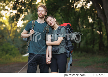 Couple wearing backpacks standing in forest. Photo of man and woman standing in forest at summer and holding hands. Man and woman wearing green t-shirts. Couple wearing backpacks standing in forest. Photo of man and woman standing in forest at summer and holding hands. Man and woman wearing green t-shirts. 111907758