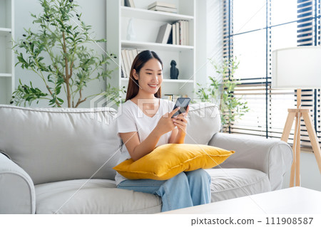 Happy young asian woman relax on comfortable couch at home texting messaging on smartphone Happy young asian woman relax on comfortable couch at home texting messaging on smartphone 111908587