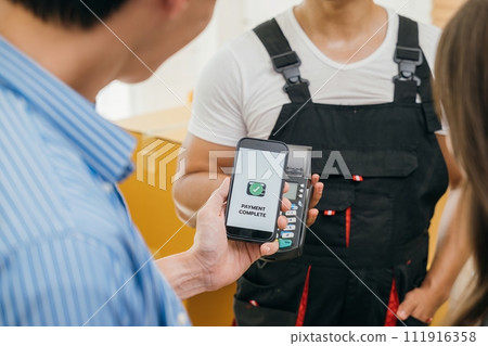 After moving couple pays via smartphone QR code. Worker carries box. Professional movers ensure smooth home relocation. Business and industry evident. Online payment for moving 111916358