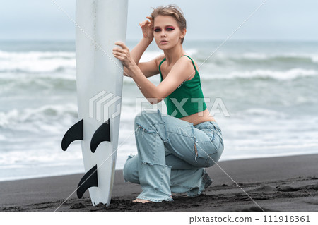 Portrait of hipster woman surfer holding surfboard vertically, sitting on beach on background of breaking sea waves. Blonde fashion model looking at camera, wearing casual clothing top and blue jeans Portrait of hipster woman surfer holding surfboard vertically, sitting on beach on background of breaking sea waves. Blonde fashion model looking at camera, wearing casual clothing top and blue jeans 111918361