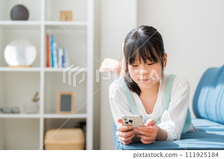 An elementary school girl operates a smartphone in the living room. 111925184