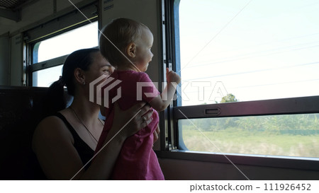 Cheerful young woman with her cute baby daughter traveling by train, looking the window with view of rural Thailand 111926452
