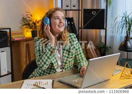 Businesswoman listening music through wireless headphones relaxing taking a break, laptop on table Businesswoman listening music through wireless headphones relaxing taking a break, laptop on table 111926968
