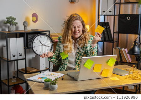 Tired businesswoman using laptop sticky notes with many tasks overworked checking time on clock Tired businesswoman using laptop sticky notes with many tasks overworked checking time on clock 111926980