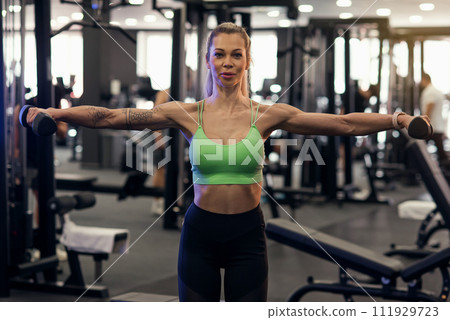 Muscular fitness woman athlete doing heavy weight exercise for shoulders with dumbbells in the gym 111929723