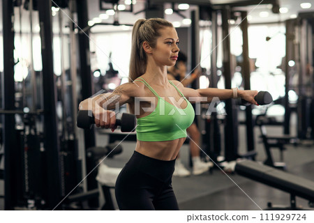 Muscular fitness woman athlete doing heavy weight exercise for shoulders with dumbbells in the gym 111929724
