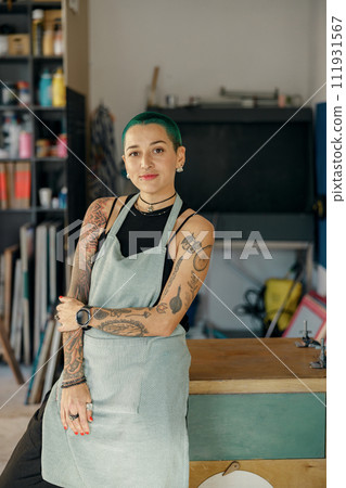 Smiling female designer standing on background of own workshop and looks camera Smiling female designer standing on background of own workshop and looks camera 111931567