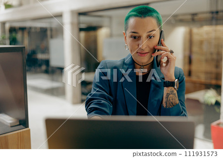 Creative smiling tattooed business woman working on laptop and talking phone in modern office Creative smiling tattooed business woman working on laptop and talking phone in modern office 111931573