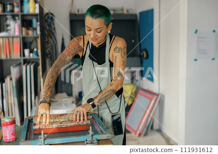 Female worker with squeegee is printing images on textile by silkscreen method in a design studio Female worker with squeegee is printing images on textile by silkscreen method in a design studio 111931612