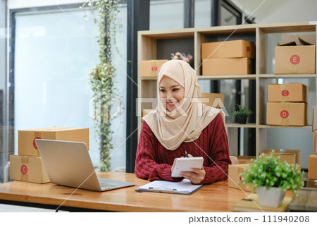 A portrait of a successful young Asian Muslim female online seller sits at her working desk with a laptop and cardboard boxes on a table. 111940208