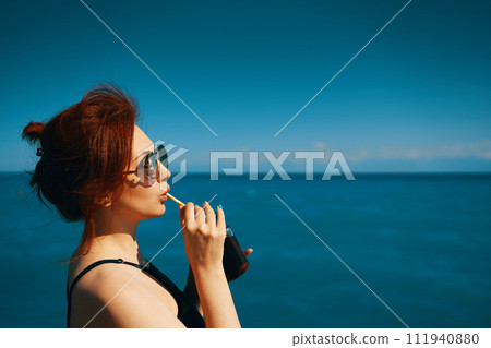 Woman on summer vacation. Red-haired girl in sunglasses drinks lemonade from bottle through cocktail tube. Blue sea, sunlight and clear sky on background. Copy space for travel agency advertising. Woman on summer vacation. Red-haired girl in sunglasses drinks lemonade from bottle through cocktail tube. Blue sea, sunlight and clear sky on background. Copy space for travel agency advertising. 111940880