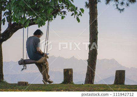 landscape and travel concept with solo freelancer man use tablet work at wooden swing under large tree and layer of mountain 111942712