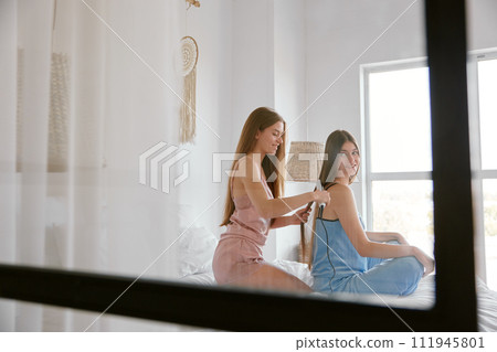 Two young female friends wearing pajamas hairstyling while spending time at home 111945801
