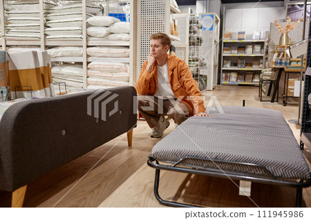 Young man customer making decision between folding bed and couch to buy Young man customer making decision between folding bed and couch to buy 111945986