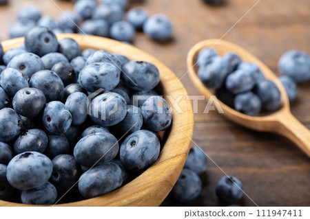 Ripe blueberries in a wooden bowl and spoon on a background of scattered berries. 111947411